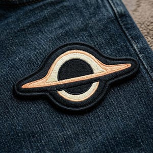 May include: Embroidered patch featuring a black hole design. The patch has a black background with a circular black center, surrounded by a cream-colored ring and a peach-colored outer edge. The patch is sewn onto a blue denim fabric.