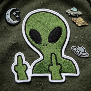 May include: Green embroidered alien patch with a white border, giving the middle finger with both hands. Also, a crescent moon and stars patch, and three pins of a UFO and planets on a green background.
