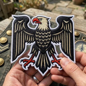 May include: Embroidered patch featuring a black eagle with outstretched wings, a red beak and talons, and a cream-colored head. The patch has a white border and is held in a person's hands. The eagle design is detailed and intricate.