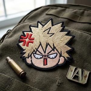 May include: Embroidered patch featuring a cartoon character with spiky blonde hair and an angry expression. The patch has a black border and a red symbol. A brass bullet casing and a metal pin with "UA" are also visible.