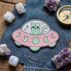 May include: Embroidered patch featuring a green cat in a pink UFO with crescent moon and star details. The patch is on a denim background, surrounded by crystals and a compass.