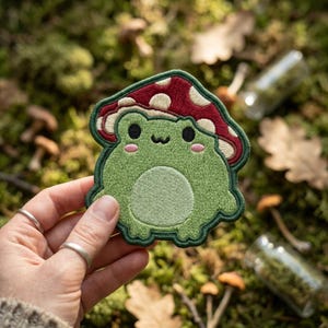May include: Embroidered patch featuring a green frog with a red mushroom cap. The frog has a smiling face with pink cheeks and black eyes. The patch is outlined in green and is held against a mossy background.