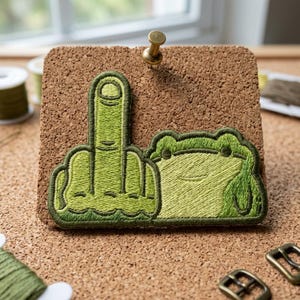 May include: Embroidered patch featuring a green frog and a hand giving the middle finger. The patch is in shades of green and is attached to a corkboard with a brass tack. The frog has a smiling face.