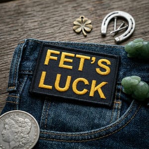 May include: Close-up of a pair of blue denim jeans with a pocket. A black patch with the words "FET'S LUCK" in yellow is sewn on the jeans. A silver horseshoe, a four-leaf clover charm, two jade rabbit figurines, and a silver coin are also visible.