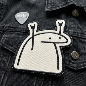 May include: A black denim jacket with a white and black embroidered patch featuring a cartoon character with raised arms. A silver guitar pick with the text "AC/DC" is pinned above the patch.
