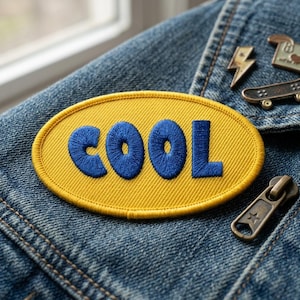May include: A yellow oval patch with the word "COOL" embroidered in blue. The patch is on a denim jacket. Also visible are a lightning bolt pin, a skateboard pin, and a zipper.
