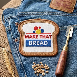 May include: Embroidered patch in the shape of a bread slice with the text "MAKE THAT BREAD" in red and blue. The patch is on blue denim jeans, next to a bread lame and wheat. The patch is approximately 3 inches wide.