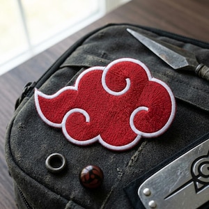 May include: Embroidered red cloud patch with white trim, resembling a stylized cloud. The patch is placed on a dark gray canvas bag, alongside a metal kunai and other accessories. The patch is a popular anime-themed accessory.