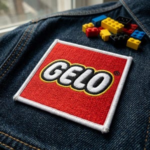 May include: A red embroidered patch with the word "GELO" in white, outlined in black and yellow, on a denim jacket. Small colorful toy building blocks are scattered nearby. The patch has a white border.