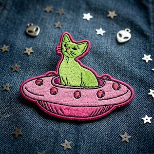 May include: Embroidered patch featuring a green cat in a pink UFO. The patch has a pink outline and is set against a blue denim background. Metallic star and alien charms are scattered around the patch.