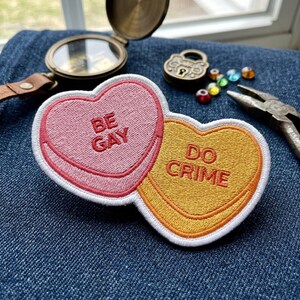 May include: Embroidered heart-shaped patches with the phrases "BE GAY" and "DO CRIME" in red lettering. The patches are pink and yellow with white and orange borders. Other items in the image include a compass, lock, pliers, and colorful beads.