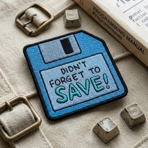 May include: A square embroidered patch shaped like a blue floppy disk with a light blue label. The text DIDN'T FORGET TO SAVE! is stitched in black and teal. The patch features a black border and a grey sliding metal tab detail.