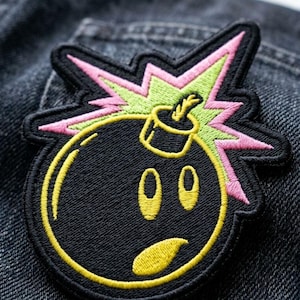 May include: Embroidered patch featuring a black bomb with a yellow face and outline. The bomb has a pink and green explosion design. The patch is on a dark denim background.