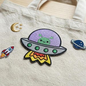 May include: A beige tote bag with embroidered patches of a purple UFO with a green alien, a rocket ship, a crescent moon with stars, and a blue planet with a ring. The patches are colorful and whimsical.