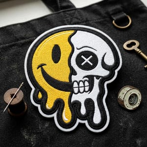 May include: Embroidered patch featuring a split design: a yellow smiley face on the left and a white skull on the right. The patch has black outlines and dripping details, with a black background.