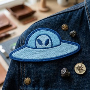 May include: A light blue embroidered patch of a UFO with an alien face, sewn onto a dark blue denim jacket. The patch is surrounded by small metal pins, including star shapes and a compass.