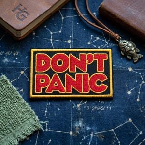 May include: Embroidered patch with the words "DON'T PANIC" in red letters on a black background, framed by a gold border. The patch is rectangular and sits on a blue fabric with a constellation pattern.