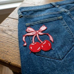 May include: Close-up of blue denim jeans with a decorative patch. The patch features a pair of red cherries with white highlights and a pink bow. The jeans have a classic five-pocket design and are made of a durable denim fabric.