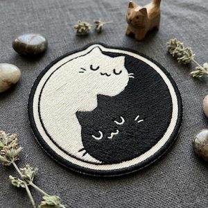 May include: A circular embroidered patch featuring a yin-yang design composed of two stylized cats. One cat is white with black embroidered details, and the other is black with white embroidered details. Both cats have closed eyes and whiskers.