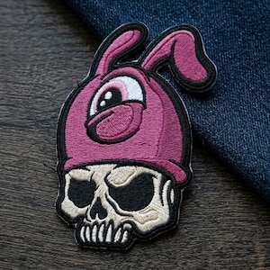 May include: Embroidered patch featuring a skull wearing a pink bunny hat. The skull is beige with black eye sockets and teeth. The bunny hat is pink with black trim. The patch is on a wooden surface with a denim background.