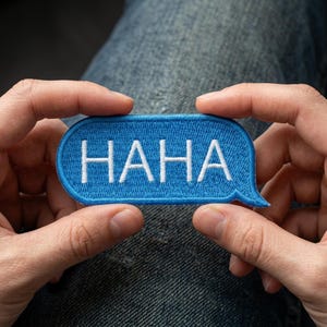 May include: A blue embroidered patch in the shape of a speech bubble, with the word "HAHA" in white capital letters. The patch is held between two hands, against a background of denim jeans.