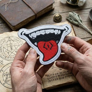 May include: Embroidered patch featuring a stylized open mouth with sharp white teeth, a black interior, and a red tongue. The patch has a white border and is held against a backdrop of aged paper and a leather-bound book.