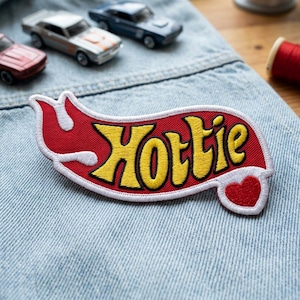 May include: Embroidered patch with the word "Hottie" in yellow, outlined in white, on a red background. A small red heart is on the right side. The patch is on a denim surface, with toy cars in the background.