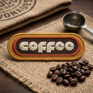 May include: An embroidered patch with the word "COFFEE" in off-white, set against a brown background with orange and yellow borders. The patch rests on burlap, with coffee beans and a metal scoop nearby. A burlap sack is in the background.