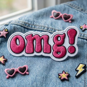May include: Embroidered patch with the word "OMG!" in pink letters with a white border, placed on a denim jacket. The patch is surrounded by small decorative patches, including pink heart-shaped sunglasses and star shapes.