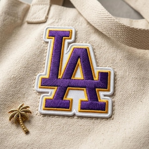 May include: Cream-colored tote bag with a large purple and gold embroidered "LA" patch. A small gold palm tree pin is attached to the bag. The bag has a matching cream-colored strap.