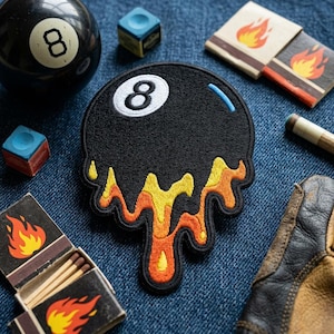 May include: A black embroidered patch shaped like a melting eight ball with orange and yellow flames. The image also includes a pool ball, chalk cubes, matchbooks with flame graphics, and a pool cue.