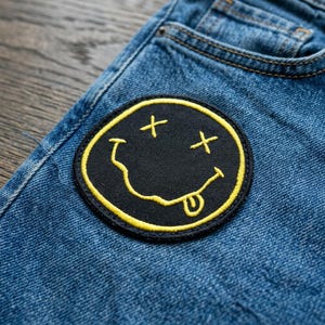May include: A black and yellow smiley face patch with crossed-out eyes and a tongue sticking out, sewn onto blue denim jeans. The patch has a yellow border and is circular in shape. The jeans have a classic five-pocket design.