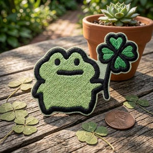 May include: Embroidered patch featuring a green frog holding a four-leaf clover. The frog has a black outline and a smiling face. The clover is green with a black outline. A penny and real clovers are next to the patch.