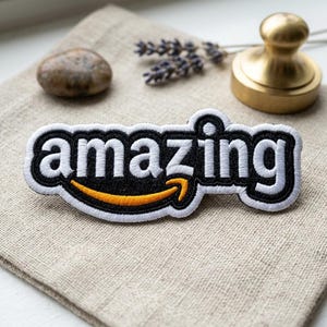 May include: Embroidered patch with the word "amazing" in white letters on a black background, outlined in white. The design includes an orange arrow. The patch is on a beige fabric surface.