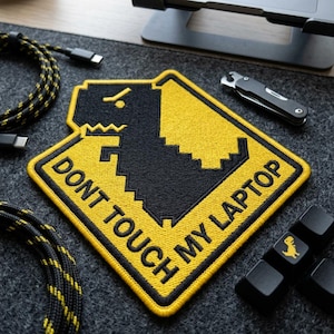 May include: A yellow and black embroidered patch with the text "DON'T TOUCH MY LAPTOP" and a dinosaur graphic. Also visible are black keyboard keys with a dinosaur icon, a black and yellow cable, and a small multi-tool.