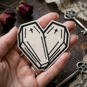 May include: Heart-shaped patch featuring two coffin designs with black outlines and small crosses. The patch is made of beige fabric and has a stitched border. The patch is held in a hand.