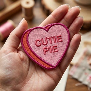 May include: A pink heart-shaped embroidered patch with the words "CUTIE PIE" in a darker pink. The patch has an orange border and a darker pink outline. The patch is held in a hand.