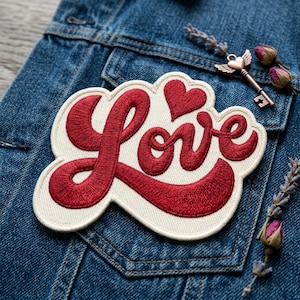 May include: An embroidered patch featuring the word Love in deep red cursive script on a cream-colored background, accented by a small red heart above the letter v. The patch is placed on a blue denim fabric next to a metal key charm and lavender.