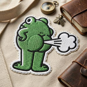 May include: Embroidered patch featuring a green cartoon frog with a white and black outline, emitting a cloud of white gas. The patch is on a beige fabric surface, with a compass, a small bronze fly, and leather-bound notebooks.