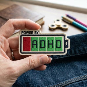 May include: Embroidered patch with the text "POWER BY ADHD" in black letters on a green battery-like design. The patch has a white border and a red section indicating low power. A lightning bolt is on the right.