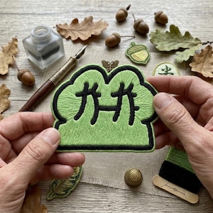 May include: A green embroidered patch with Japanese characters, outlined in black, held by hands. The patch is surrounded by acorns, leaves, a thimble, and a vintage pen and ink set. The patch has a cloud-like shape.