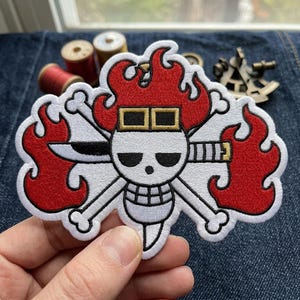May include: An embroidered patch featuring a white skull with black eye sockets and a grid-patterned jaw, wearing a pair of goggles on its forehead. The skull is surrounded by red flames and crossed white bones, with a horizontal sword behind it.