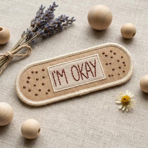 May include: A tan fabric patch in the shape of a bandage with the embroidered words "I'M OKAY" in burgundy thread. The patch has a white border and small brown stitches. Dried lavender, wooden beads, and a daisy are also in the image.