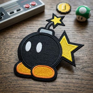 May include: Black embroidered patch of a cartoon bomb with a yellow starburst design. The patch has two white oval eyes and orange and yellow feet. A retro video game controller and other game-themed items are in the background.
