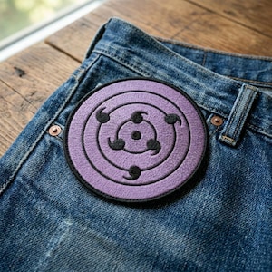May include: A round, purple embroidered patch with a black border and a spiral design, featuring black comma-shaped details. The patch is placed on a pair of blue denim jeans, showcasing a casual style.