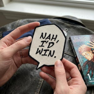 May include: A cream-colored patch with black embroidered text that reads "NAH, I'D WIN." The patch is in a speech bubble shape. A book titled "Jujutsu Kaisen" is visible in the background.