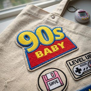 May include: Beige canvas tote bag with a large embroidered patch that reads "90s BABY" in yellow and red. Additional patches include a floppy disk and a video game controller. A vintage Tamagotchi keychain is also visible.