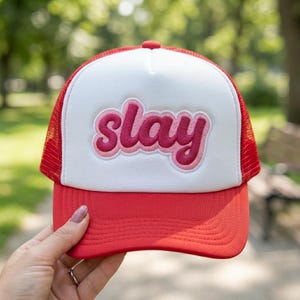 May include: Red and white trucker hat with the word "slay" embroidered in pink. The hat has a red brim and mesh backing, and a white front panel. A stylish accessory for casual wear.