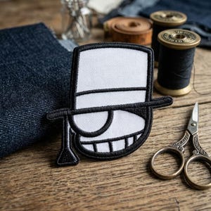 May include: A black and white embroidered patch featuring a stylized character wearing a top hat and a monocle. The design includes bold black outlines, a white hat crown, and a white monocle lens, creating a graphic, high-contrast appearance.