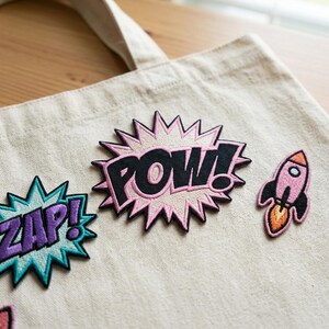 May include: A beige canvas tote bag with three embroidered patches. The patches feature the words "ZAP!" in purple and teal, "POW!" in black and pink, and a pink rocket with orange flames.